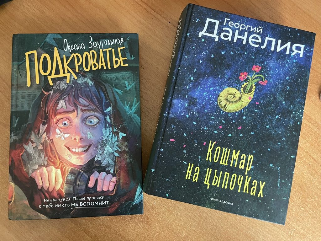 На изображении две книги на деревянном столе. Левая книга с названием 'Подкроватье' Оксаны Заугольной, изображает персонажа с яркими глазами и улыбкой, окруженного цветами. Правая книга с названием 'Кошмар на цыпочках' Георгия Данелия, имеет темный фон с изображением ракушки и цветов.